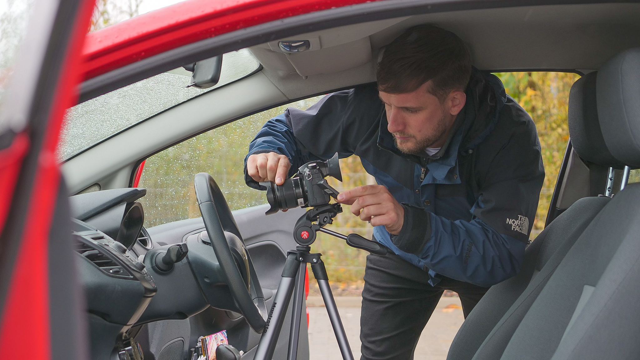 A man operating a large camera in the drivers seat of a car.